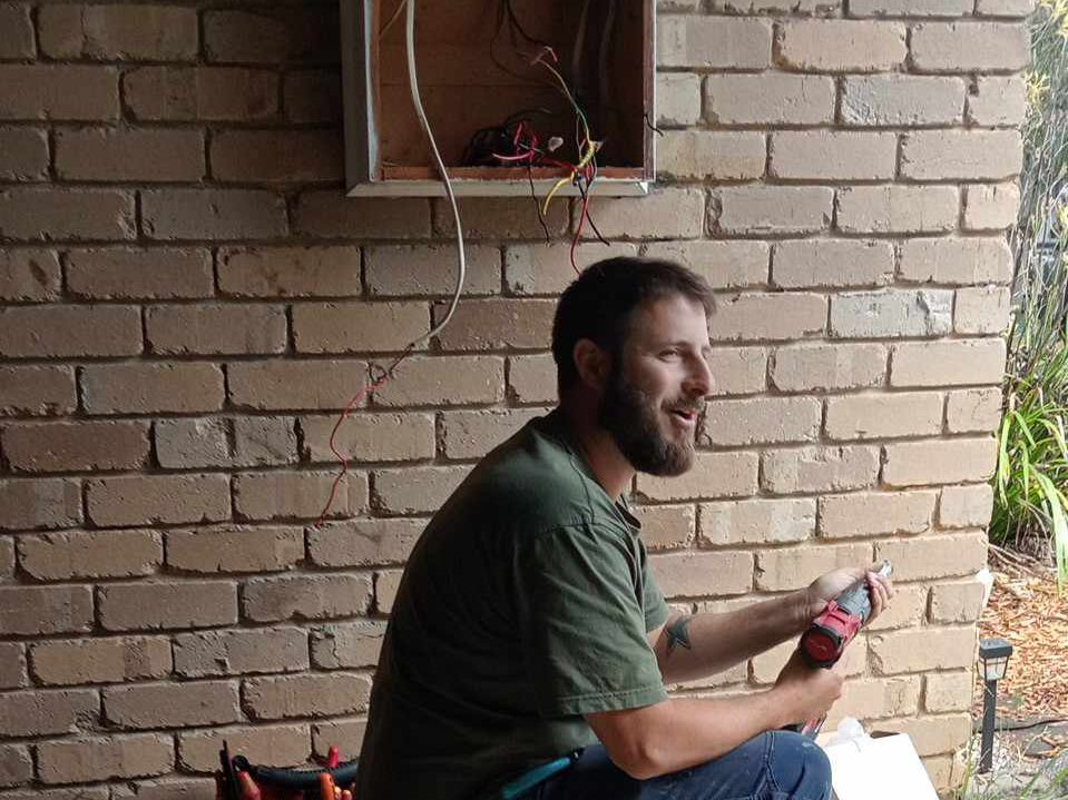 Electrician kneeling beside an open switchboard cavity on a brick wall, holding a cordless tool