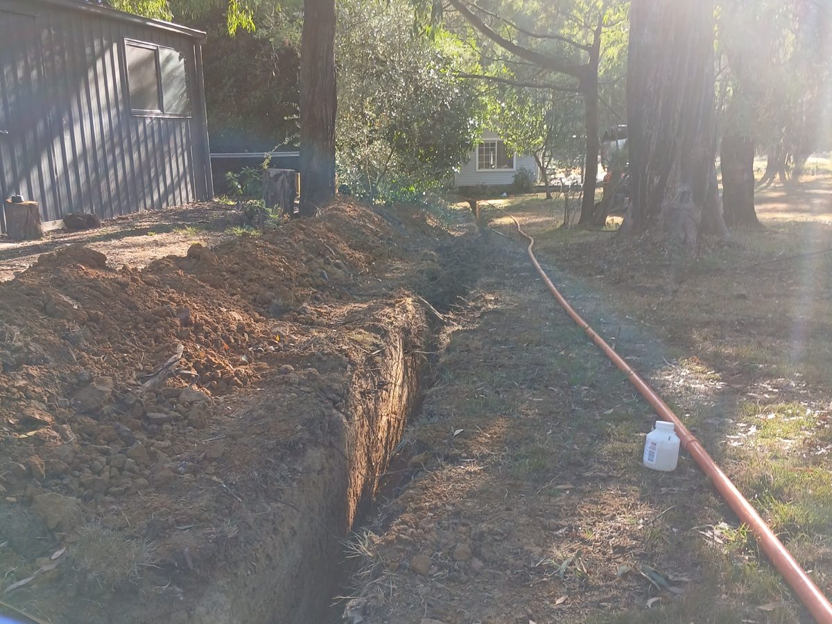 Open trench beside a shed with orange conduit laid out on the ground