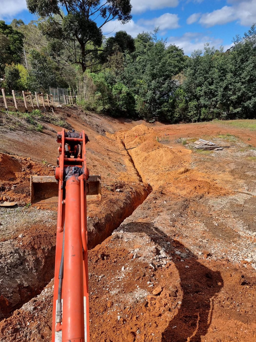 Long straight trench cut across a bush block with an excavator arm in the foreground