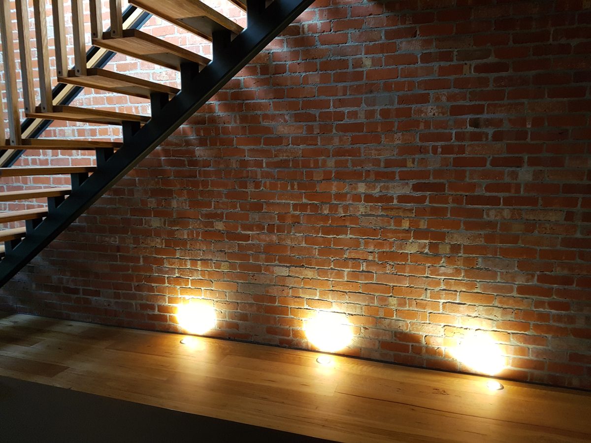 Three in-floor uplights washing light up a brick wall beside a stair