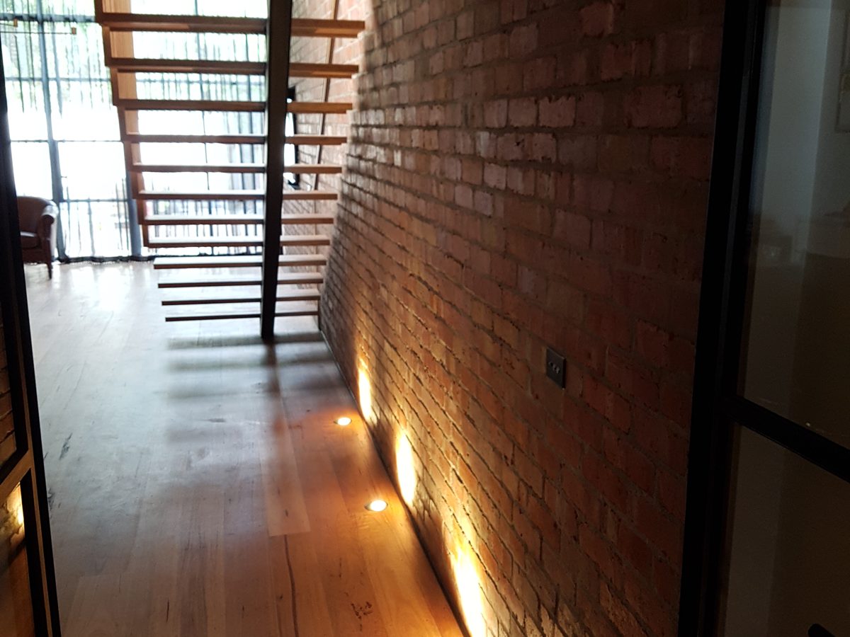 Row of in-floor uplights along a brick wall in a hallway, with a floating stair behind